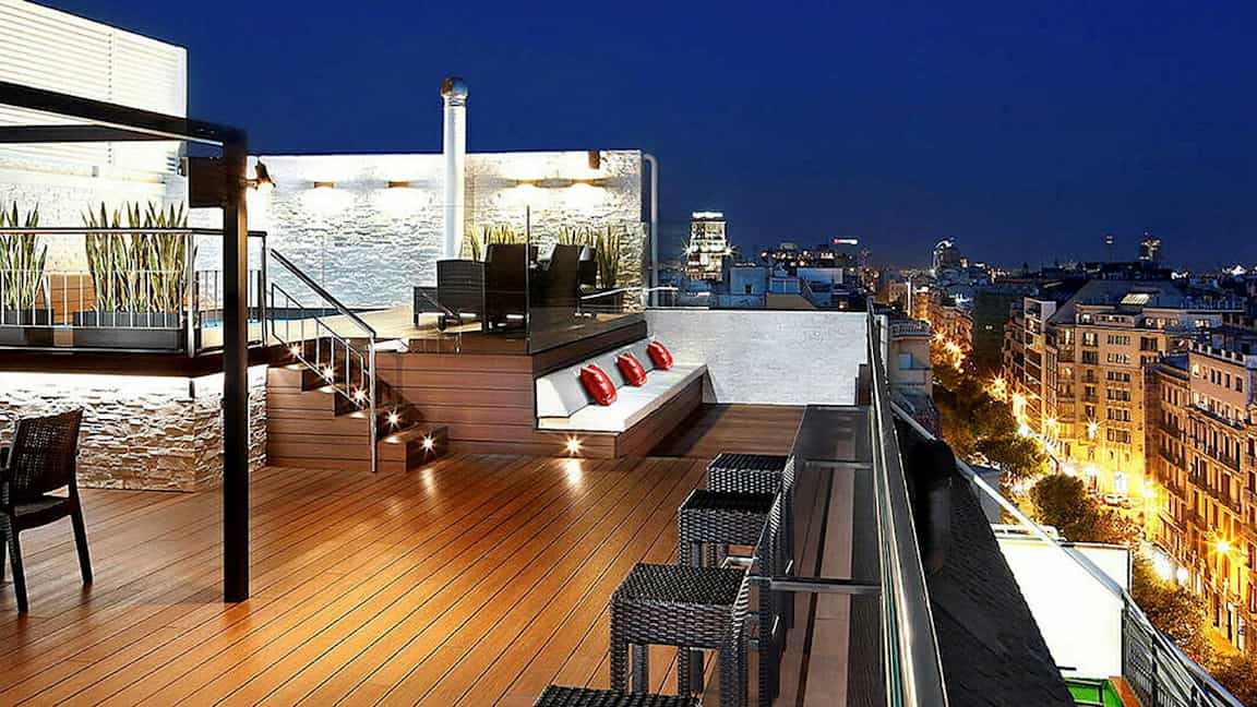 En takterrass med belysning, möbler för utomhusservering och planteringar, med utsikt över en stad på kvällen. (AI-genererad text)