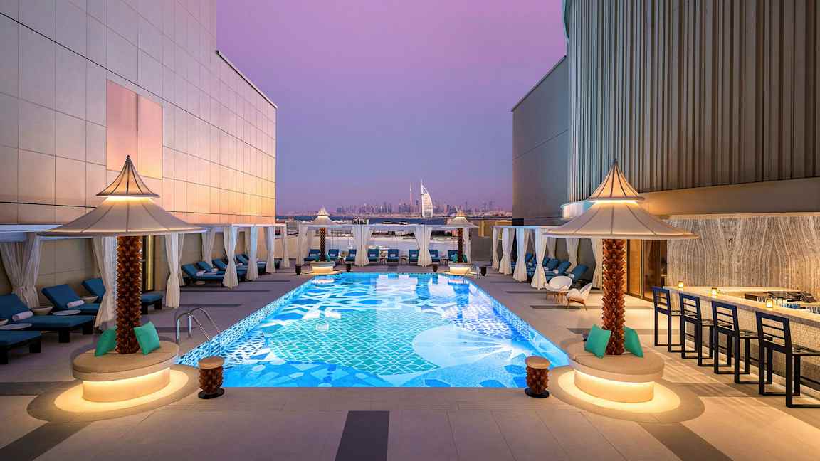 En takterrass med en dekorativ pool omgiven av solstolar och små parasoller. I bakgrunden syns Burj Al Arab och Dubais stadssilhuett under en lila kvällshimmel. (AI-genererad text)
