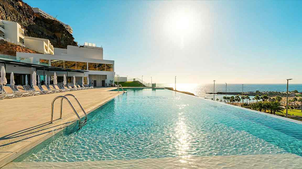 En infinitypool med spegelklart vatten, solsängar och parasoller på sidan, i ett hotellområde med utsikt mot havet och stranden. (AI-genererad text)