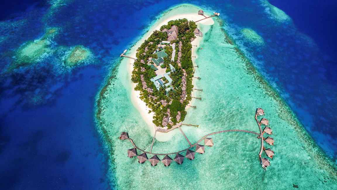 Flygbild av en tropisk ö med byggnader omgivna av turkost vatten. (AI-genererad text)