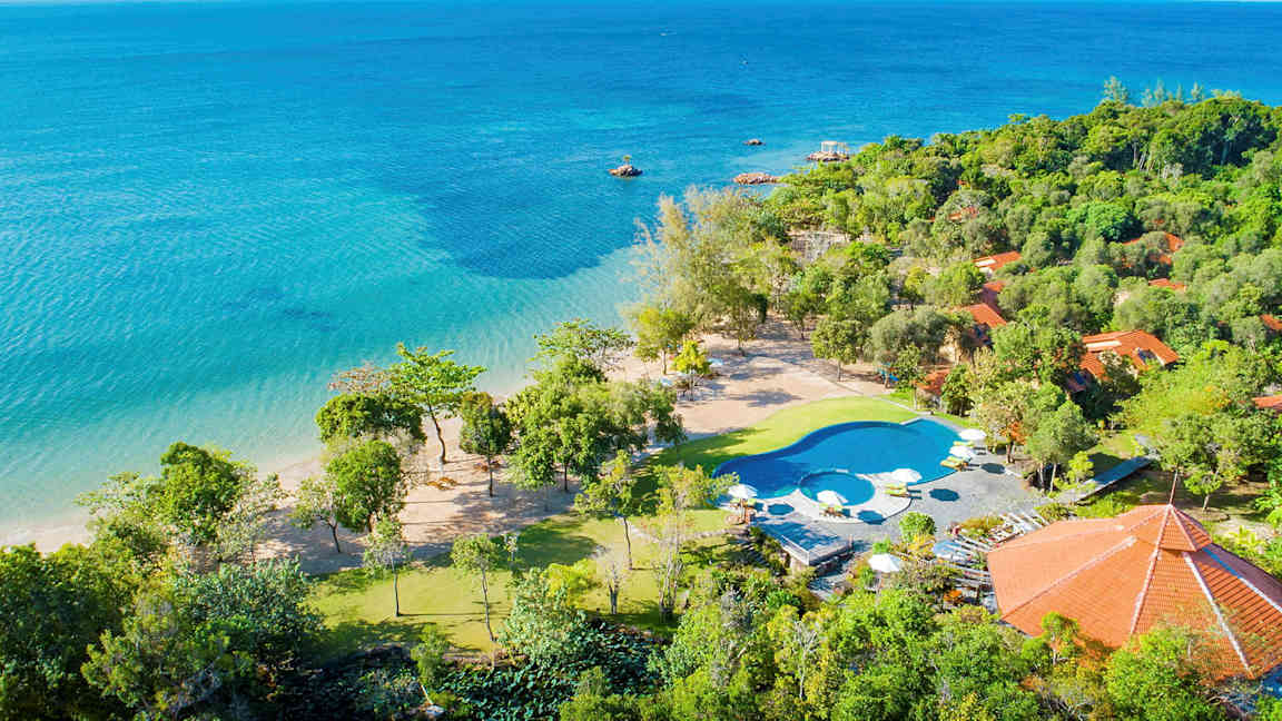 Vy över en tropisk strand med klart blått vatten, omgiven av grönska och byggnader med röda tak. En pool med solstolar är placerad nära stranden. (AI-genererad text)
