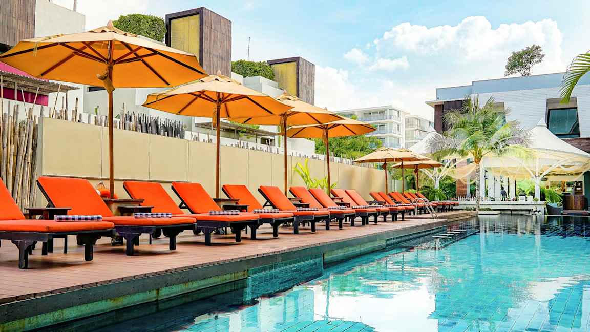 En utomhusbassäng med orangea solstolar och parasoller vid kanten, omgiven av modern arkitektur. (AI-genererad text)
