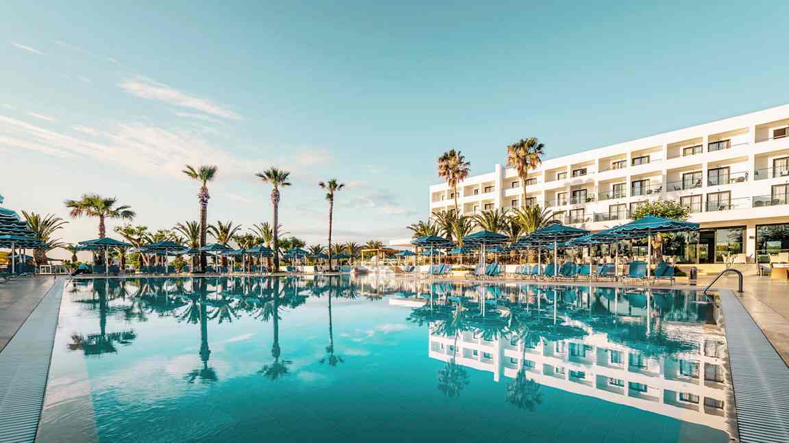 Exteriör av ett hotell med en stor pool framför och omgivande palmer. (AI-genererad text)