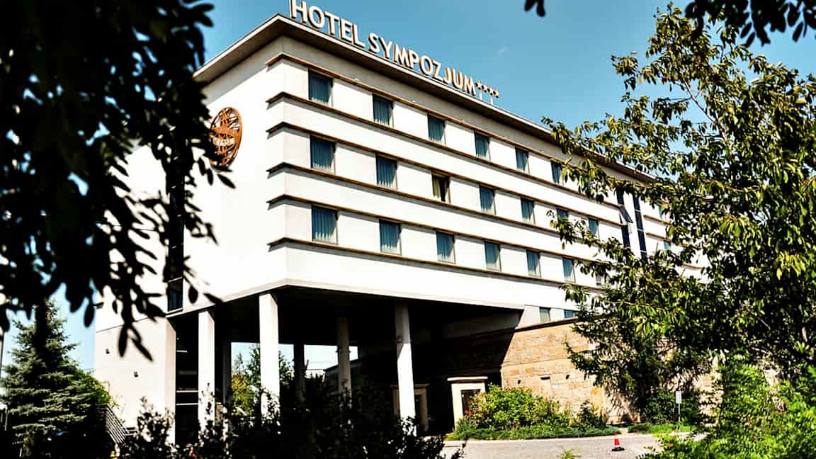 Byggnad med flera våningar, märkt med texten 'Hotel Sympozjum', omgiven av träd och växtlighet. (AI-genererad text)