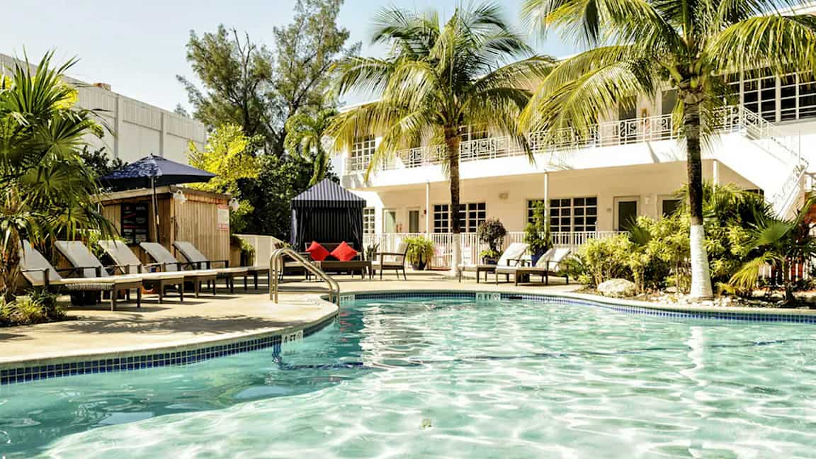 En utomhuspool omgiven av stolar, palmträd och en byggnad i bakgrunden. (AI-genererad text)