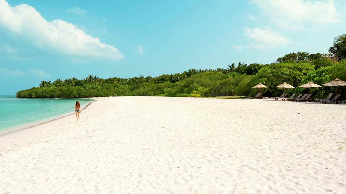 En kvinna går på en sandstrand med turkost vatten och grön vegetation i bakgrunden. (AI-genererad text)