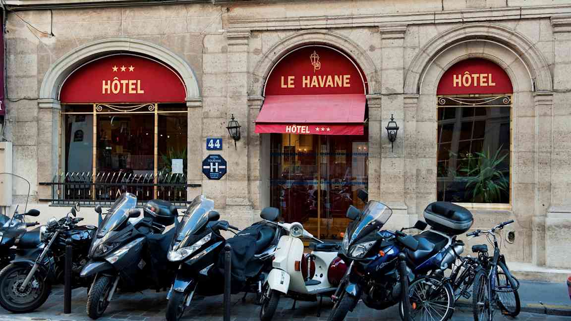 Bild som visar entrén till ett hotell med namnet Le Havane, tre stjärnor visas på markisen. Framför byggnaden står mopeder och cyklar parkerade. (AI-genererad text)