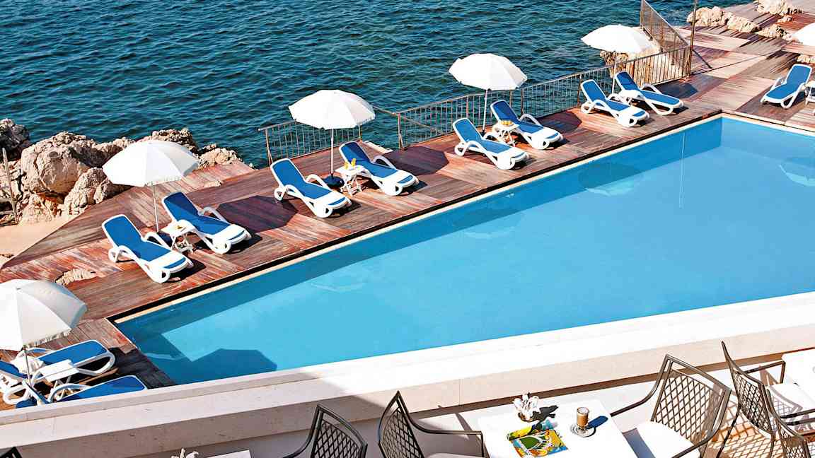 En pool med solstolar och parasoller vid en träterrass intill havet. Förgrunden visar ett bord med stolar och drycker. (AI-genererad text)