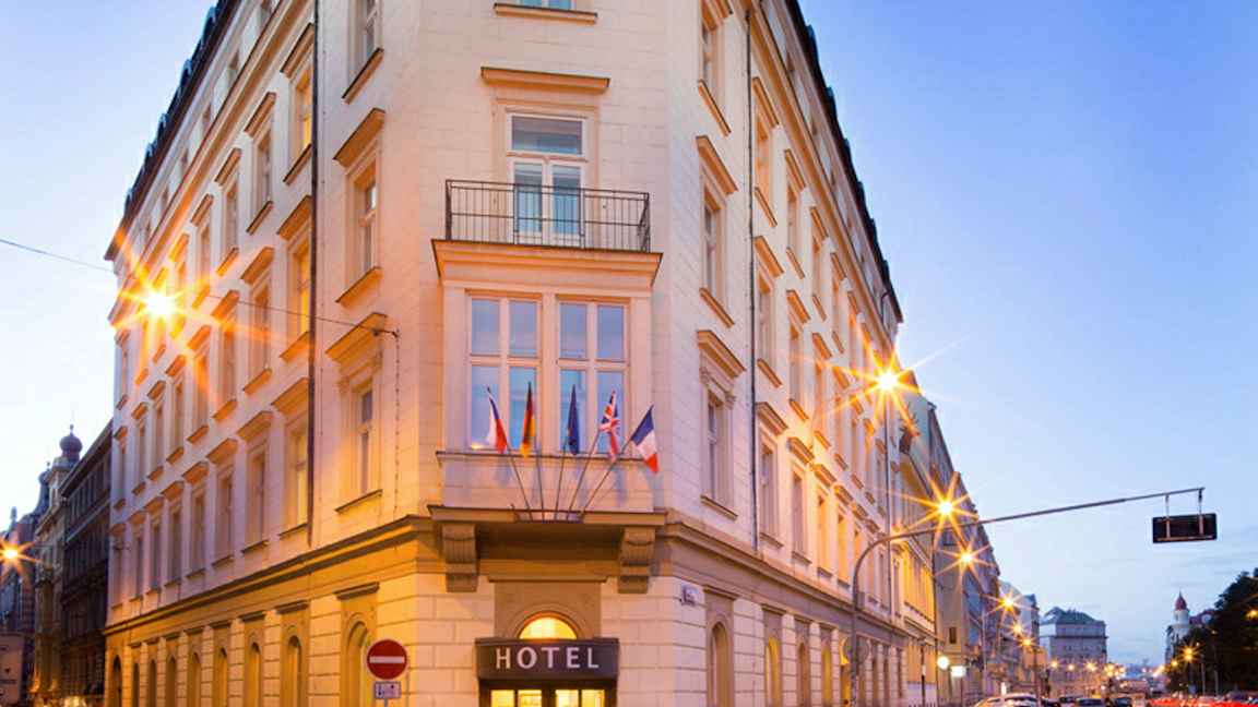 Ett hörn av en byggnad som är ett hotell, med fasad av ljus puts och dekoration. Flera flaggor hänger ovanför ingången. (AI-genererad text)