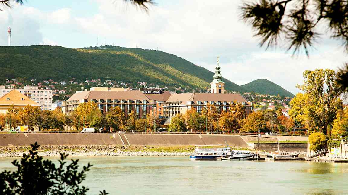 Vy över en flod med båtar, en stadsbebyggelse med en kyrka och gröna kullar i bakgrunden. (AI-genererad text)