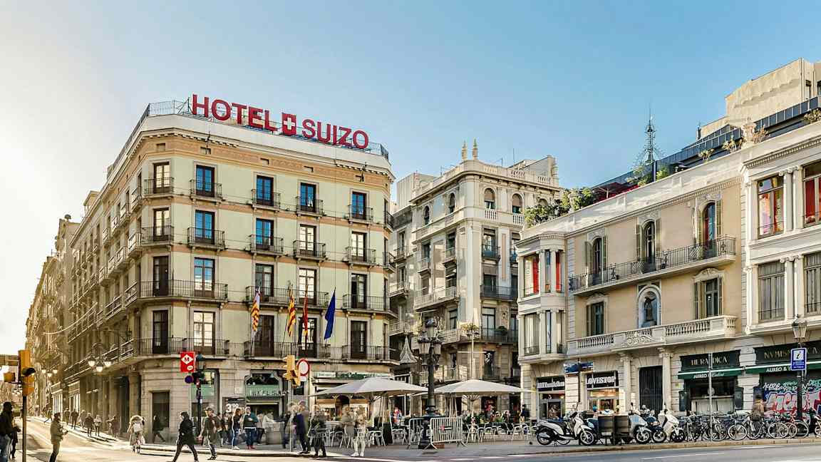 Ett gatuhörn med byggnader varav en är märkt som Hotel Suizo. Några människor går och sitter vid uteserveringar. (AI-genererad text)