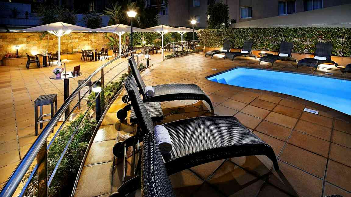 Hotellets poolområde med solstolar och parasoll vid ett upplyst och kaklat uteområde. (AI-genererad text)