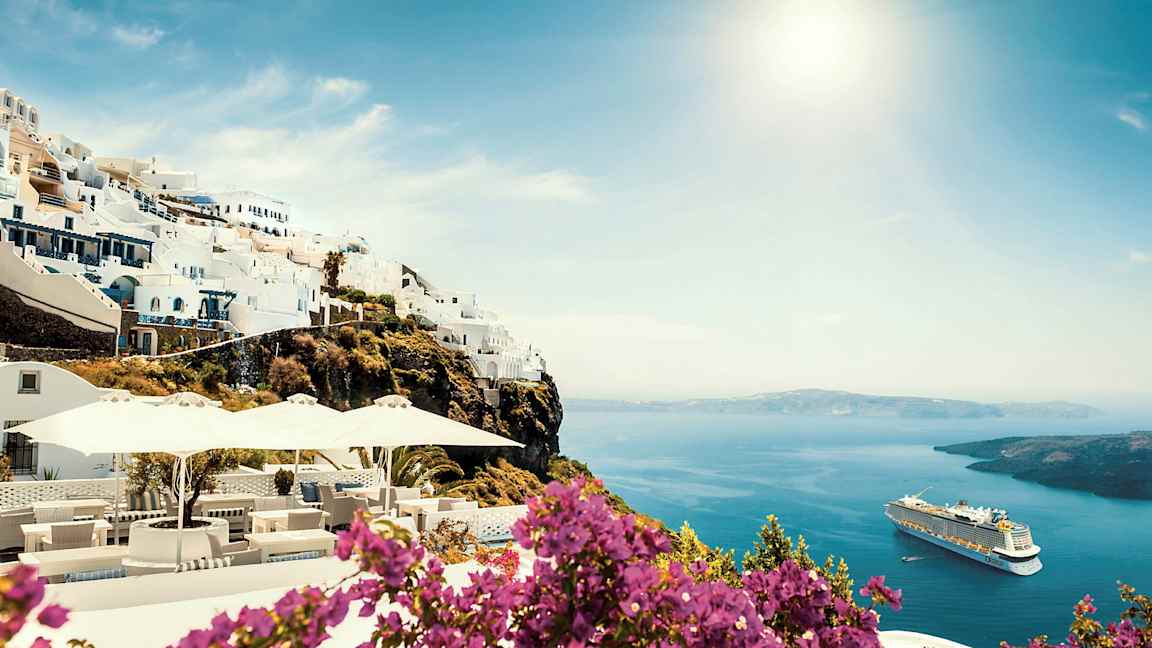 Vita byggnader på en klippa med havsutsikt och blommor i förgrunden. Ett kryssningsfartyg syns på vattnet. (AI-genererad text)