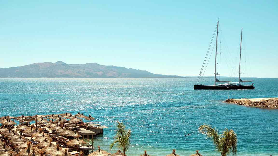 Strand med parasoller och lång segelbåt på havet med berg i bakgrunden. (AI-genererad text)