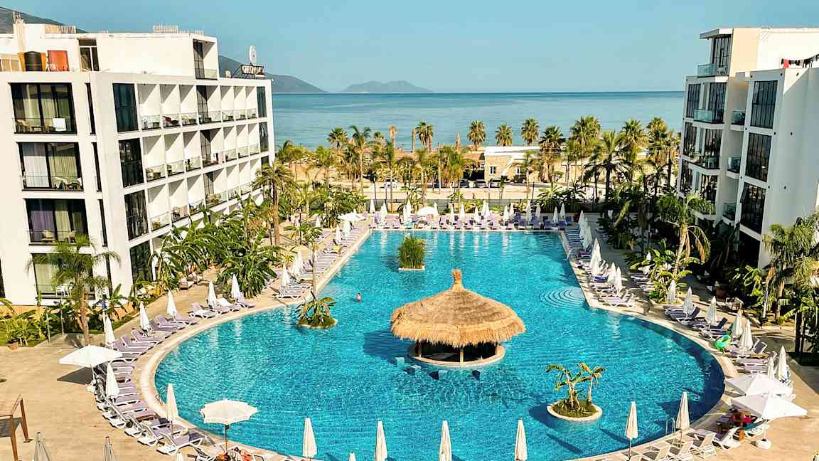 Hotellkomplex med stor pool omgiven av solstolar och parasoller, med havet i bakgrunden. (AI-genererad text)