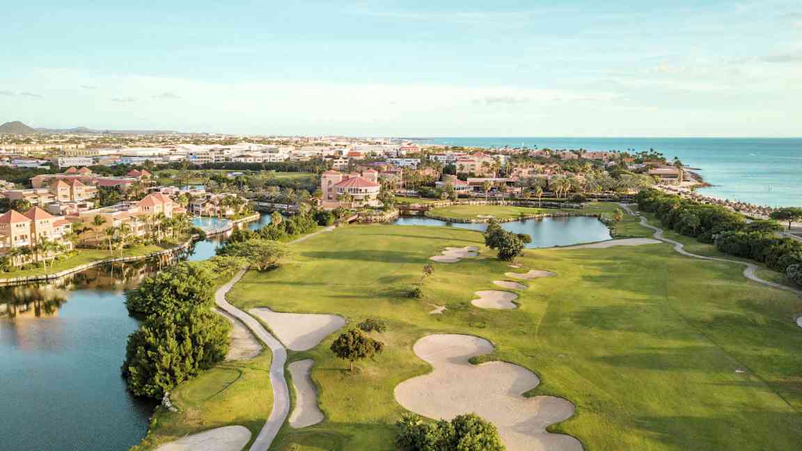 Golfbana med sandbunkrar och gröna gräsytor i närheten av vattendrag och strand. Bebyggelse i bakgrunden. (AI-genererad text)