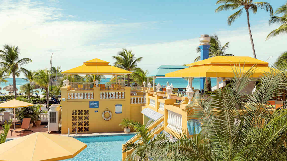 En tropisk miljö med en pool och gula parasoller, omgiven av palmer med havet i bakgrunden. (AI-genererad text)