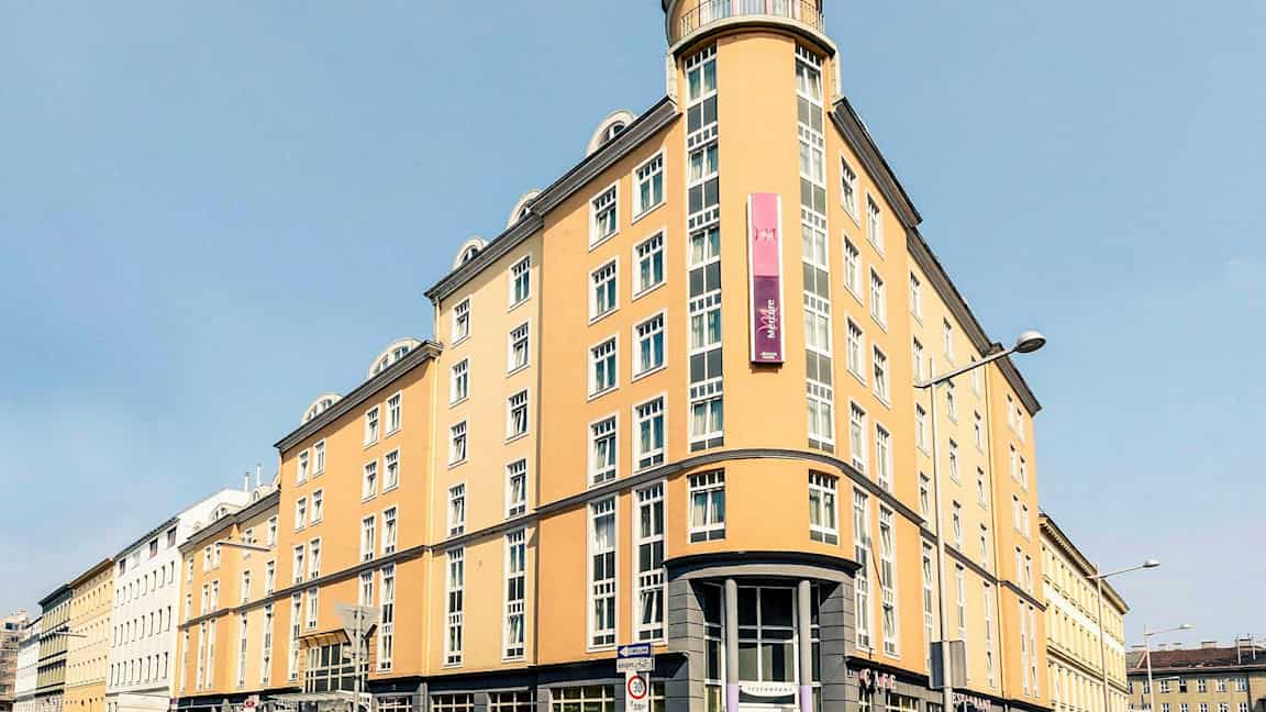 En ljusgul flerfaldigt fönstrad byggnad med skylt och rundad hörningsfasad. (AI-genererad text)