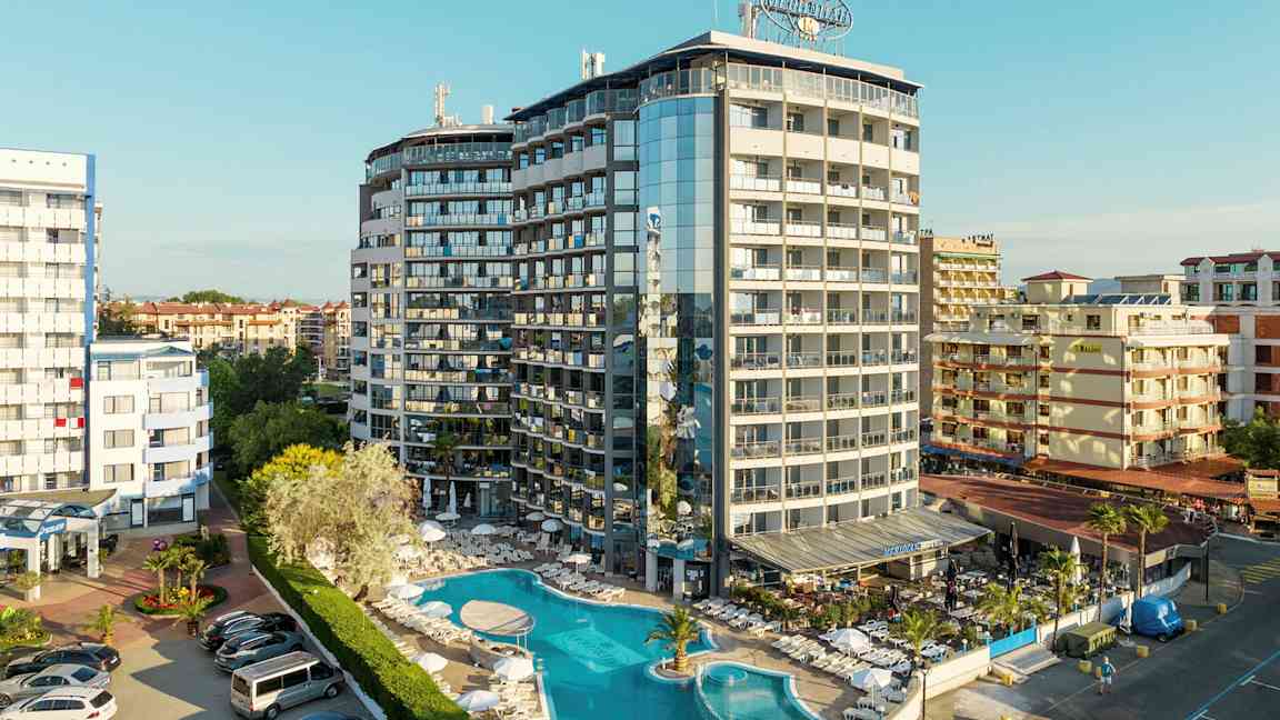 Ett flervåningshotell med pool och solstolar framför, inbäddat i ett tätbebyggt område. (AI-genererad text)