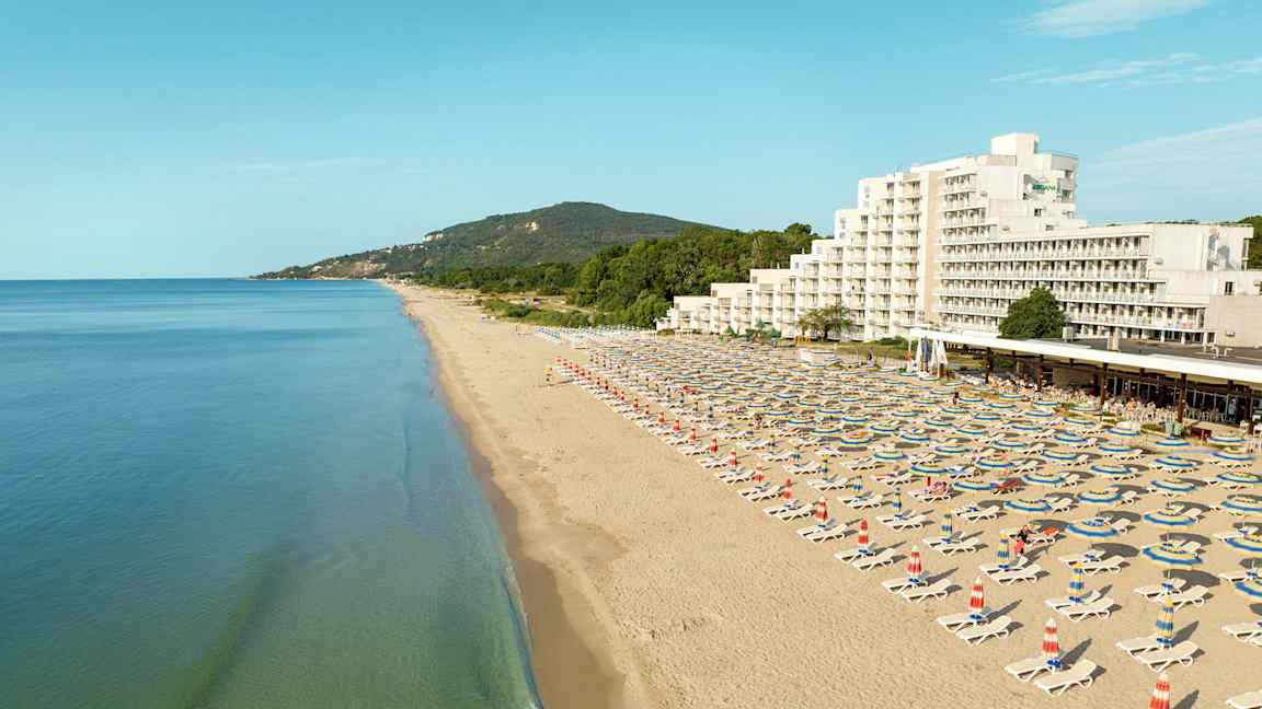En lång sandstrand med solstolar och parasoller, samt ett hotell och gröna kullar i bakgrunden. (AI-genererad text)