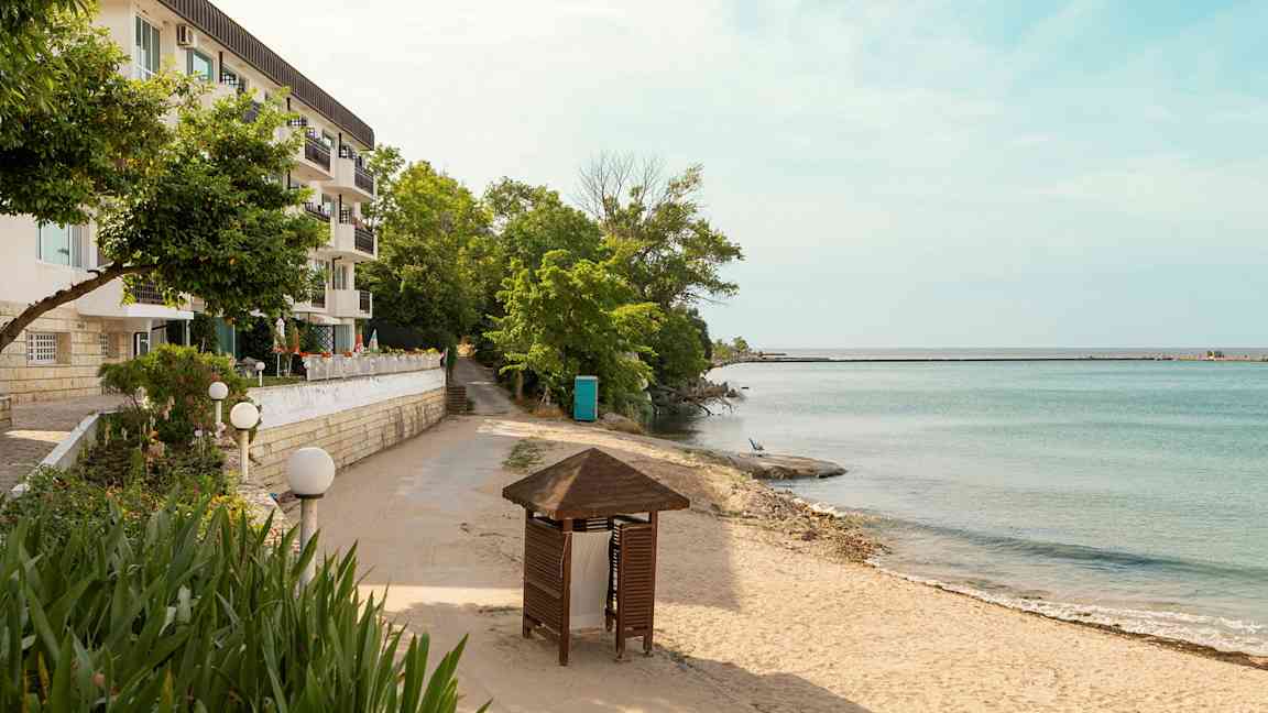 En byggnad med flera balkonger bredvid en sandstrand och hav med träd och en paviljong i förgrunden. (AI-genererad text)