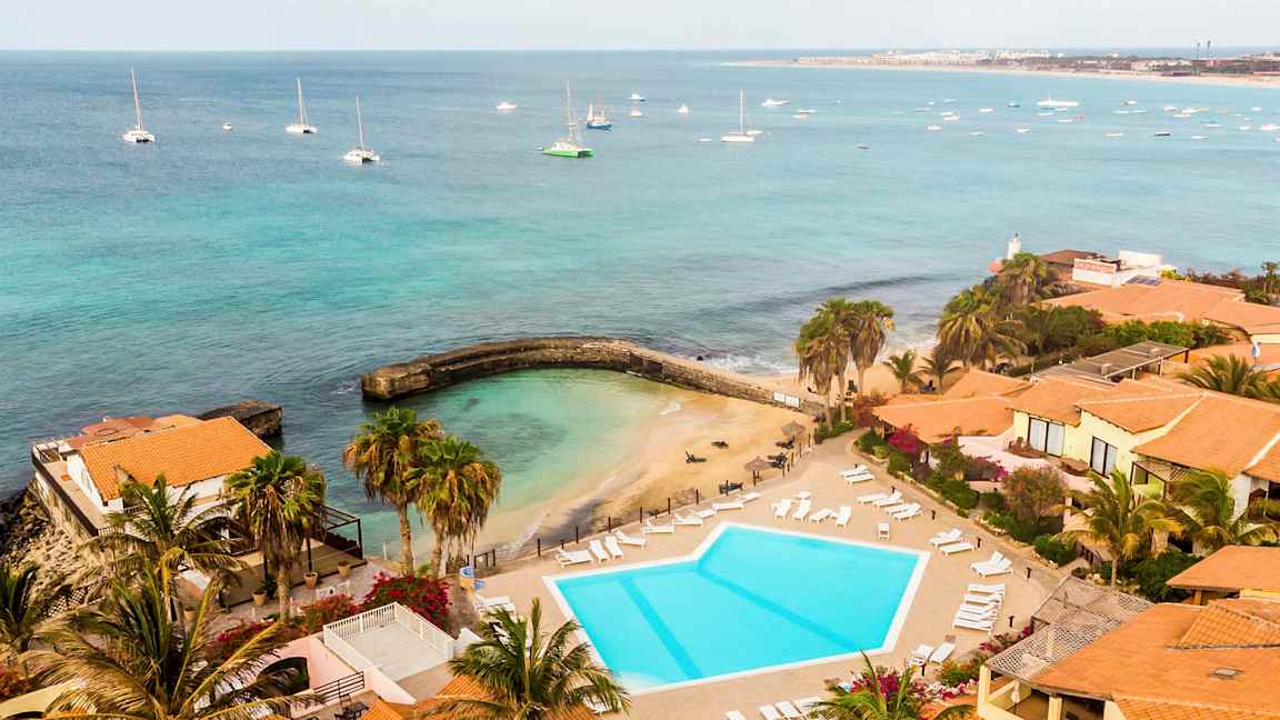 Vy över en badplats och en pool vid havet, omgiven av byggnader med tegelröda tak. (AI-genererad text)