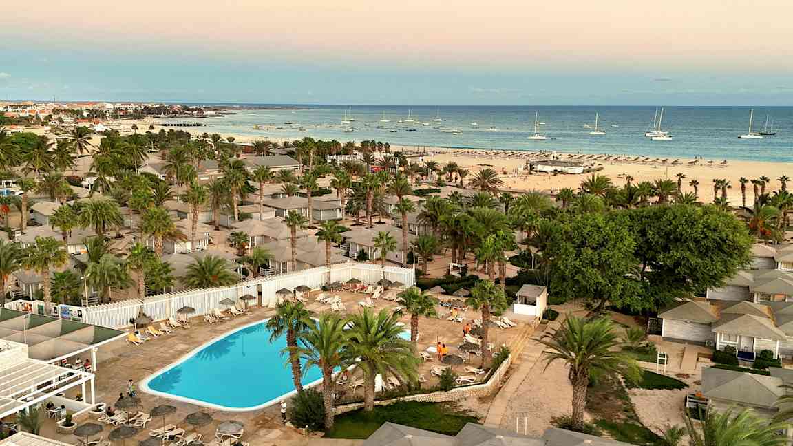 Utsikt över en resort med swimmingpool, omgiven av palmer och nära en strand med båtar i havet. (AI-genererad text)