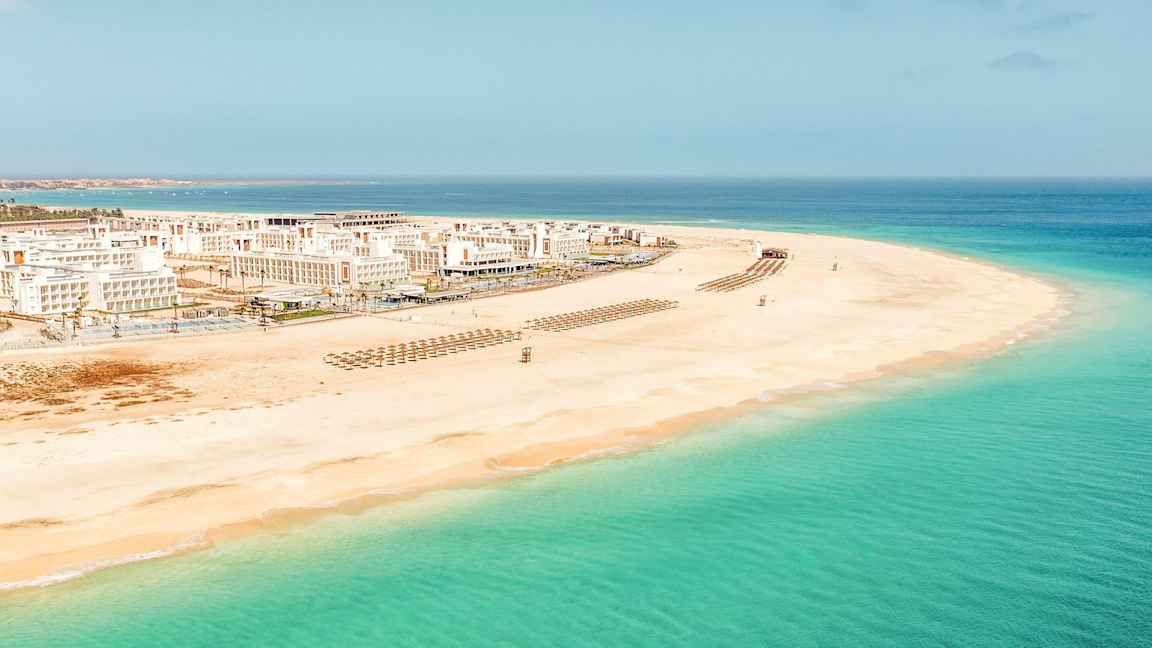 Flygfoto av en vit sandstrand med klart turkost vatten och vita byggnader i bakgrunden. (AI-genererad text)