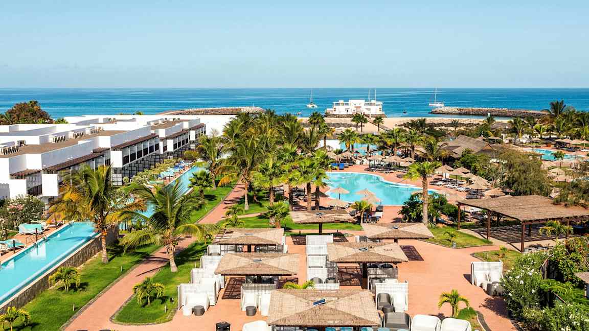 Semesteranläggning med vita byggnader, en swimmingpool och omgivande grönområde, samt hav i bakgrunden. (AI-genererad text)