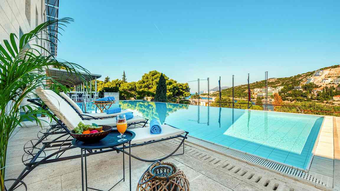 En utsikt mot en infinitypool med solstolar, bord med frukt och dryck intill sig, samt gröna omgivningar och himmel i bakgrunden. (AI-genererad text)