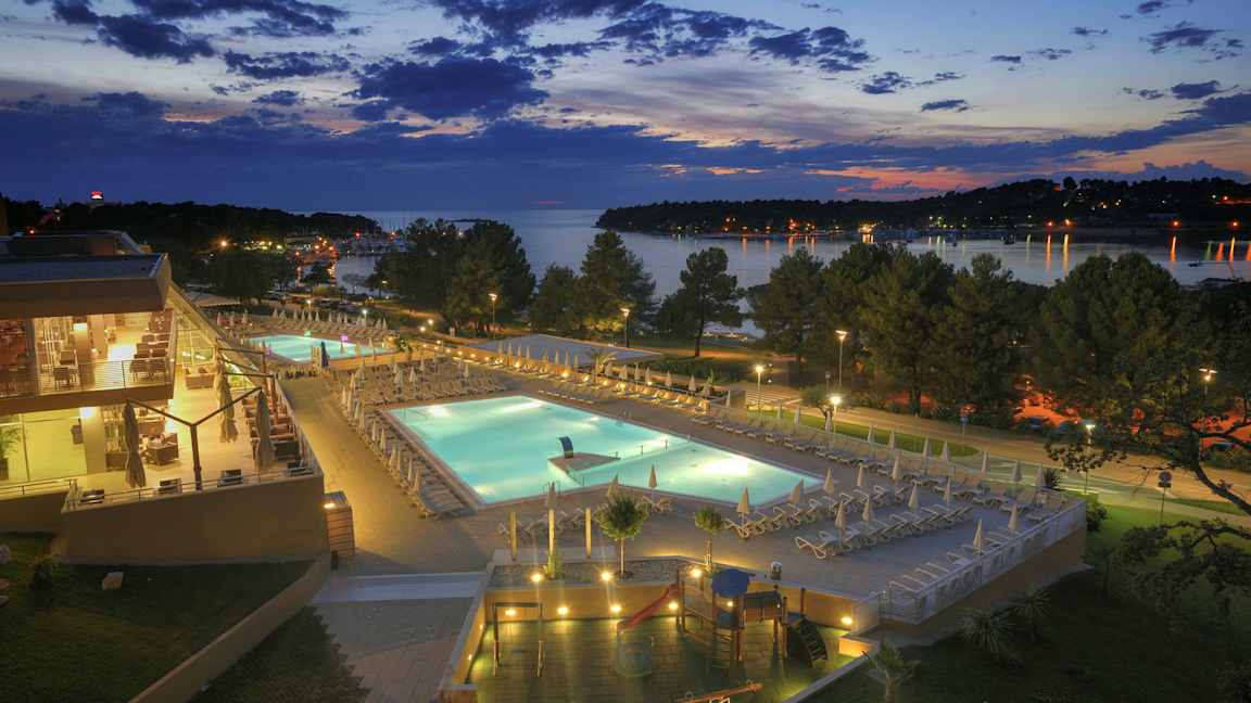 En kvällsutsikt över ett hotellområde med upplysta swimmingpooler, omgivande terrasser och träd, med en sjö i bakgrunden. (AI-genererad text)