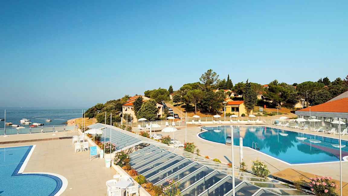 En vy över en utomhuspool omgiven av en terrass med solstolar och parasoller. I bakgrunden syns havet samt byggnader och vegetation. (AI-genererad text)