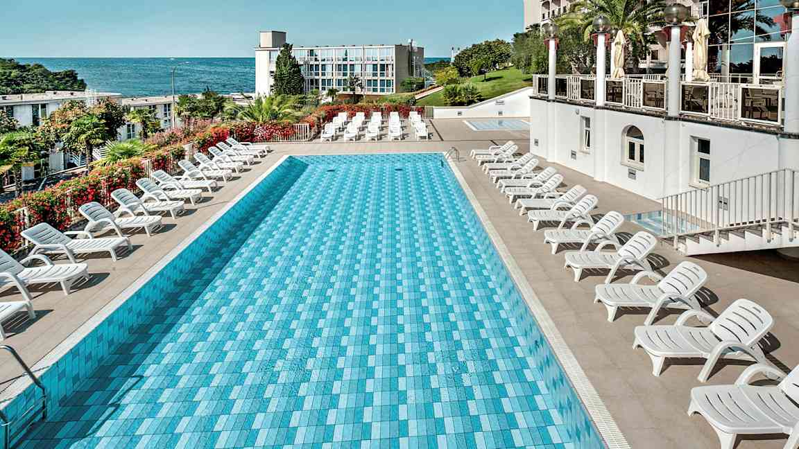 En rektangulär pool omgiven av solstolar, med havet och en byggnad i bakgrunden. (AI-genererad text)
