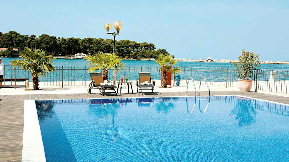 En pool med klarblått vatten och två solstolar bredvid. Havet syns i bakgrunden. (AI-genererad text)