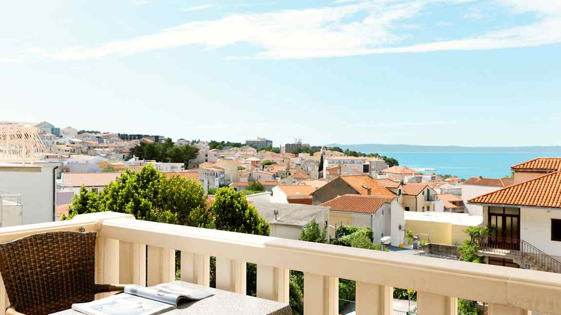 Balkong med bord och stol, utsikt över en stad med röda tak och havet i bakgrunden. (AI-genererad text)