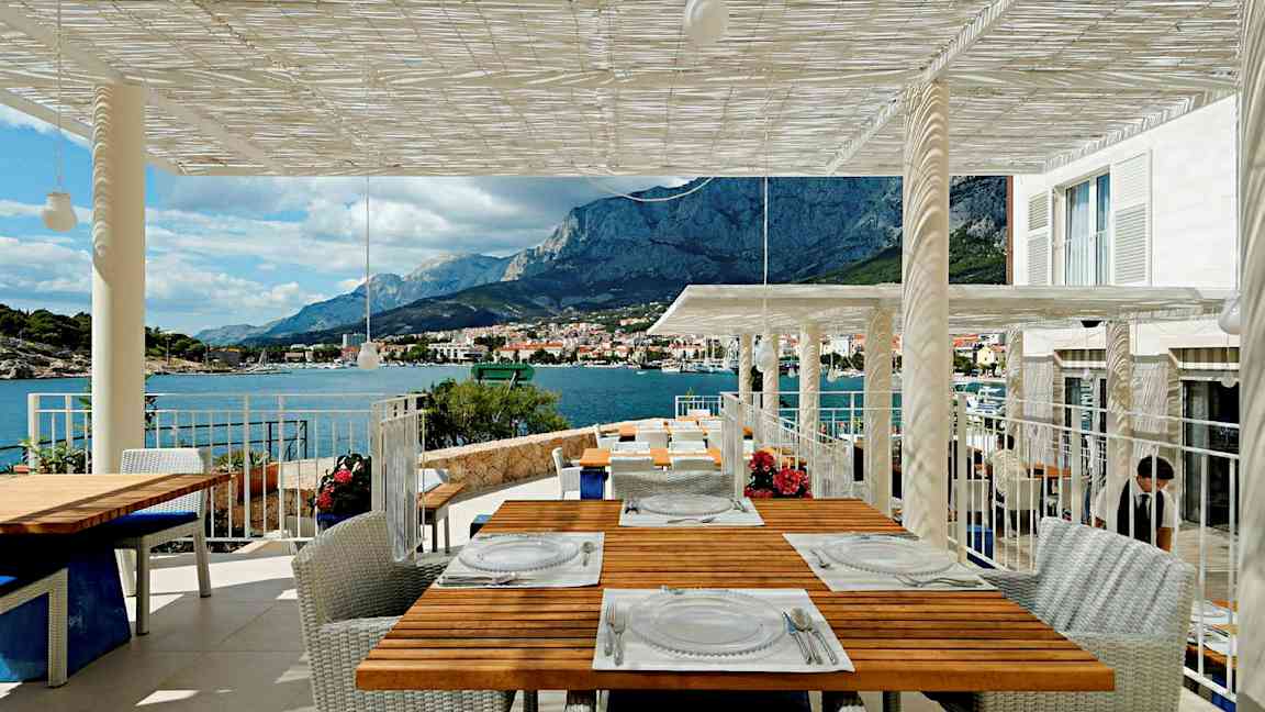 En terrass med utsikt mot berg och hav, flera bord är dukade för matgäster. (AI-genererad text)