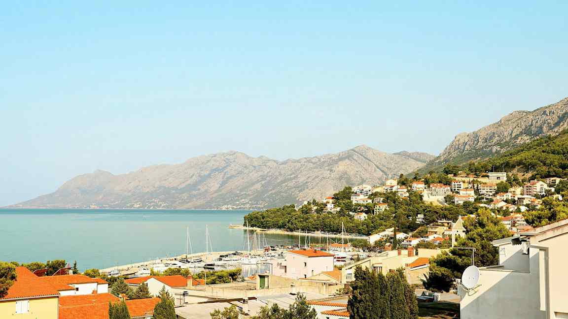 Vy över en kuststad med röda tak, en småbåtshamn och berg i bakgrunden. (AI-genererad text)