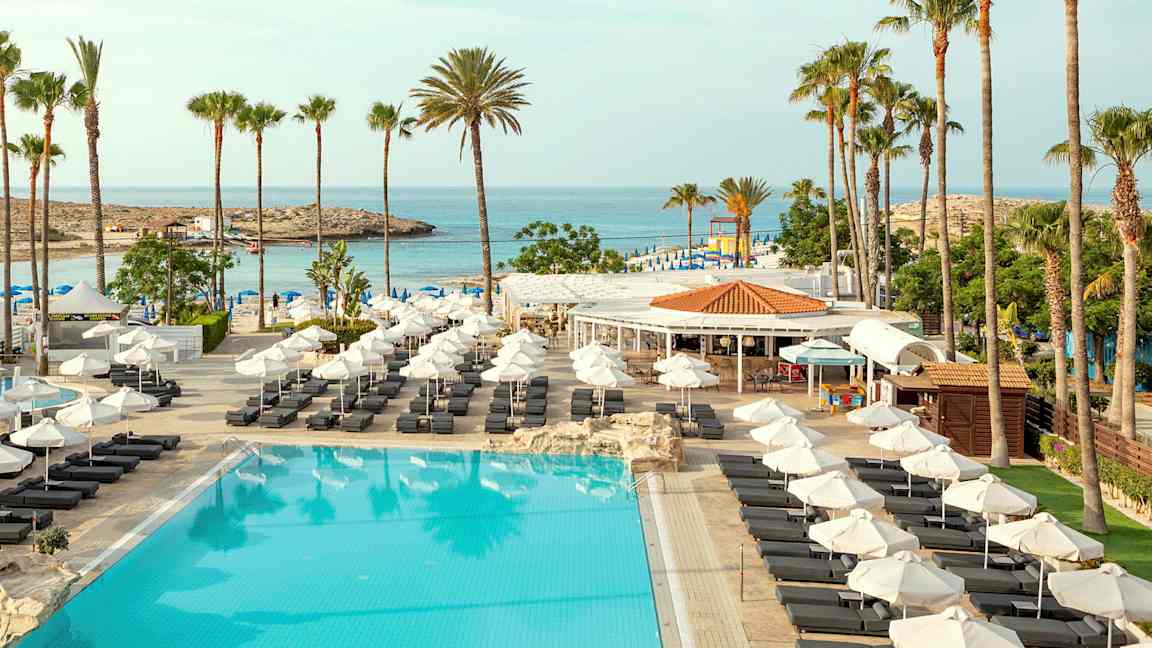 Bild av en resort med en stor pool, solstolar och palmer. I bakgrunden syns havet och en strand. (AI-genererad text)