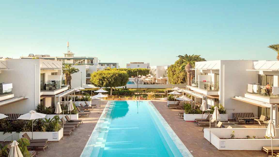 En utomhuspool omgiven av moderna vita byggnader med balkonger och solstolar under parasoll. (AI-genererad text)