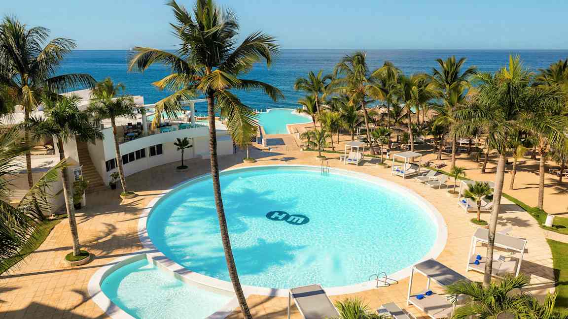 Ett hotellområde med en stor rund pool omgiven av palmer, solsängar och cabanas. Utsikt över havet i bakgrunden. (AI-genererad text)