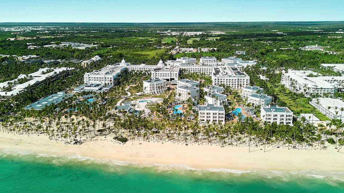 Flygbild av ett hotellkomplex vid en strand inklusive poolområde, omgiven av grönska. (AI-genererad text)