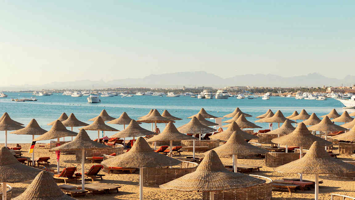 En strand med solstolar och parasoller av naturmaterial, med hav och båtar i bakgrunden. (AI-genererad text)