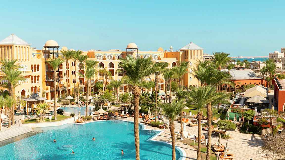 En bild på ett hotellområde med en stor pool, omgiven av palmer och byggnader i varma färger. (AI-genererad text)