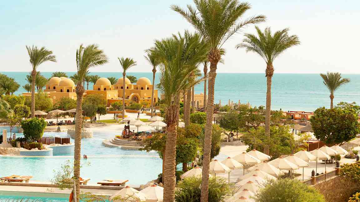 Utsikt över en poolområde med parasoller, palmer och en gul byggnad med kupoler nära havet. (AI-genererad text)