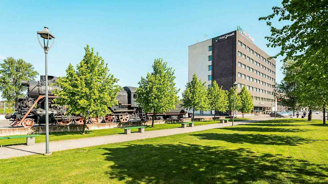 Ett gammalt ånglok står utomhus bredvid en modern hotellanläggning och grönskande träd. (AI-genererad text)