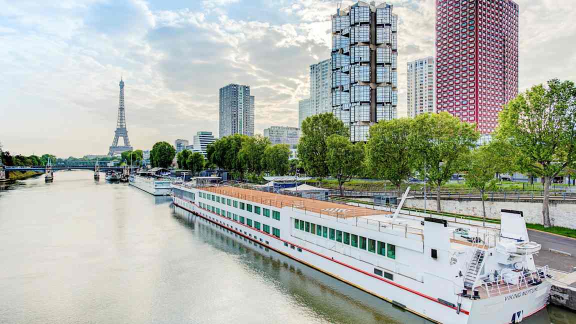 Flod med en båt vid kaj, moderna byggnader och Eiffeltornet i bakgrunden. (AI-genererad text)