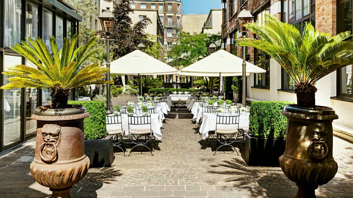 Utomhus sällskapsområde med bord och parasoller omgiven av byggnader och palmer. (AI-genererad text)