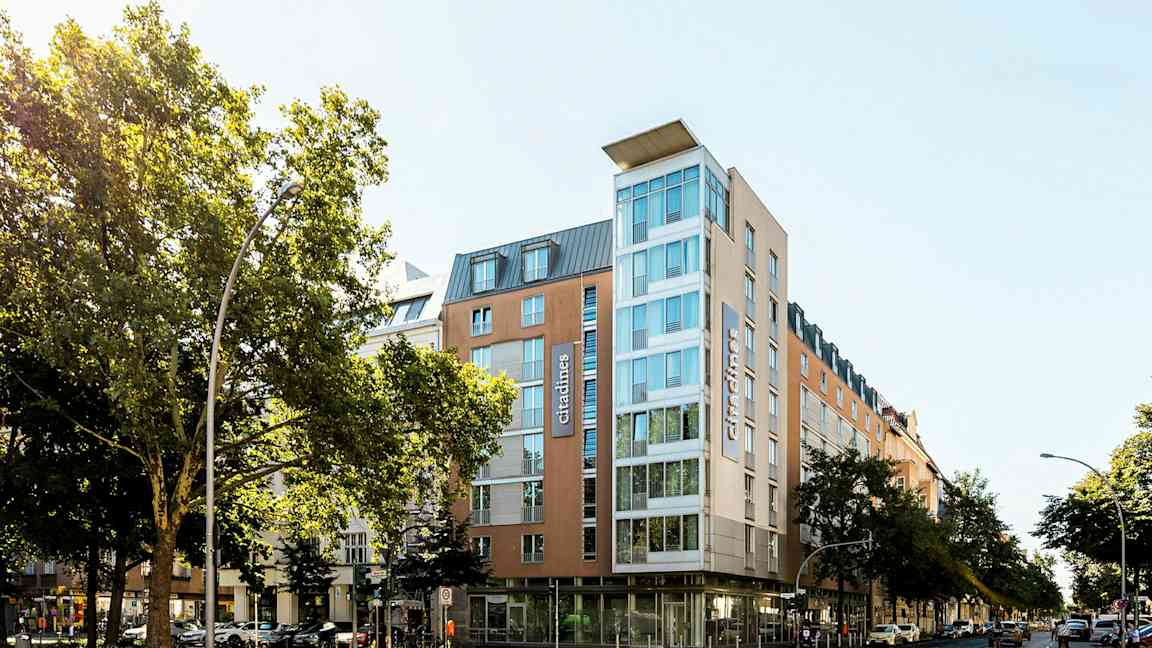 Ett modernt hotell med glasfasad omgiven av träd och stadsmiljö. (AI-genererad text)