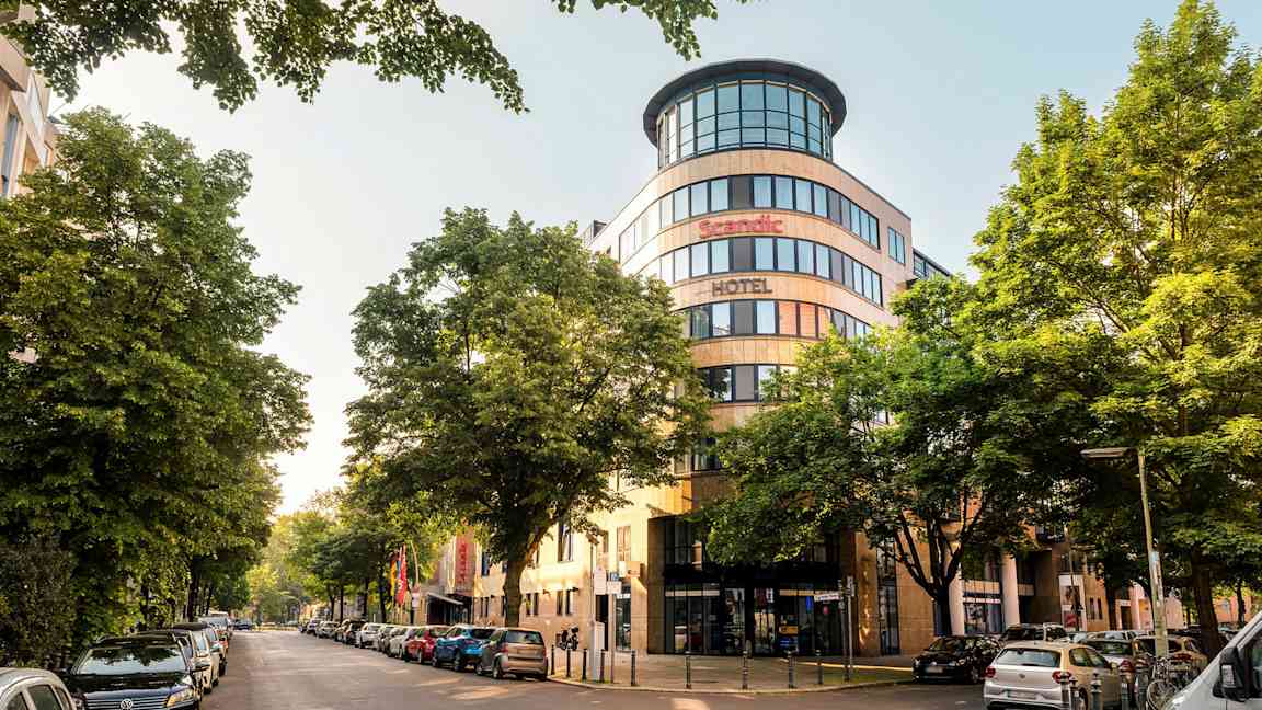 En stadsbild med fasaden av ett Scandic hotell omgiven av träd och parkerade bilar. (AI-genererad text)