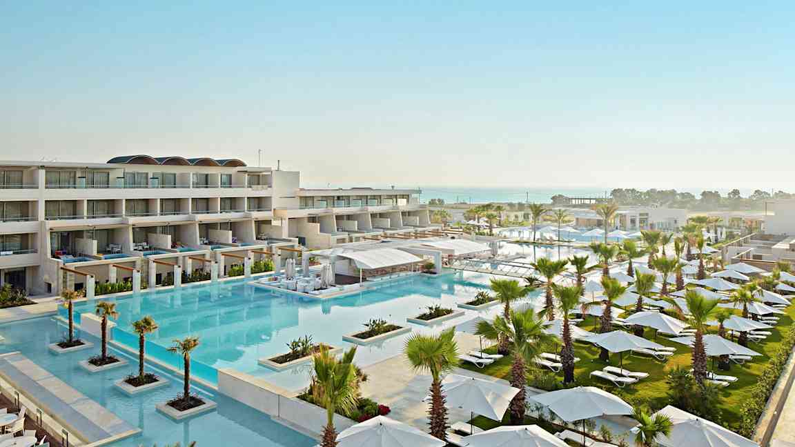 Ett hotellområde med flera byggnader, en stor pool och omgivande gröna områden fyllda med parasoller och solstolar. I bakgrunden syns havet. (AI-genererad text)